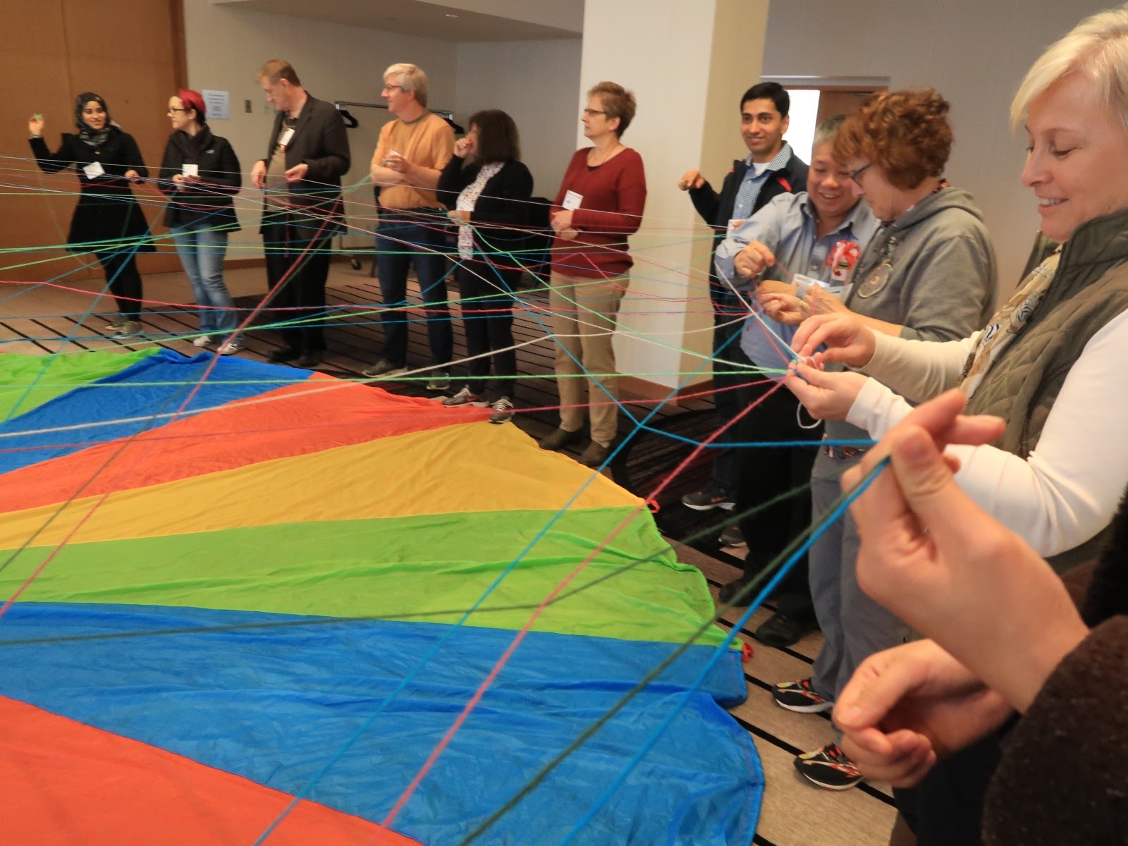 A view of part of a multi-colord parachute on the floor, surrounded by a group of people with strands of yarn wrapped around the fingers of one of their hands. Yarn strands stretch across the parachute connecting strands between those who have have made a connection with at PLoP.
