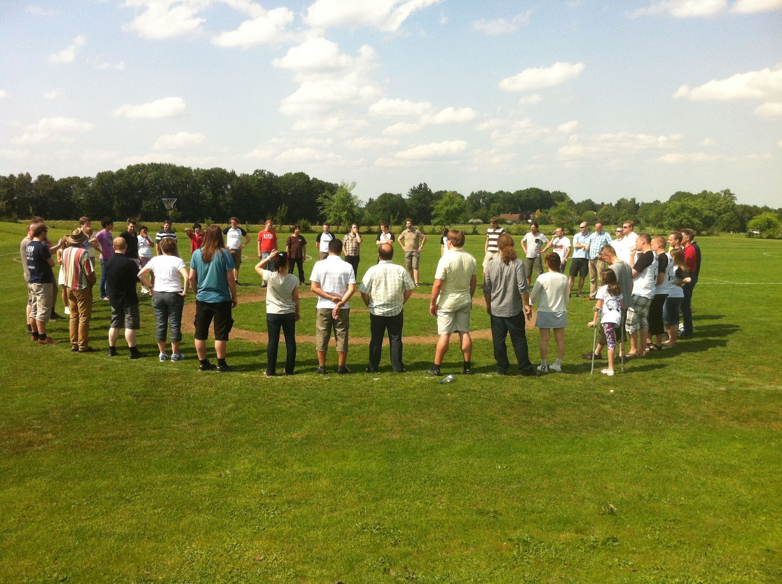People in a large circle in a field of green grass. The sky is blue with light puffy clouds. Green trees are in the background.